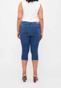 Jeans capri in denim blu scuro con vita media, due tasche posteriori e orlo pulito. Indossati con un top bianco senza maniche e tacchi con fantasia.