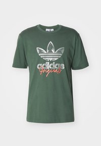 Zelené bavlněné tričko s krátkým rukávem, na přední straně s stříbrným logem Adidas a nápisem "Originals" v oranžovém kurzívě.