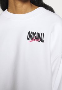 White sweatshirt featuring a black "ORIGINAL" graphic with pink accents, crew neckline, and soft fabric texture. Casual design.