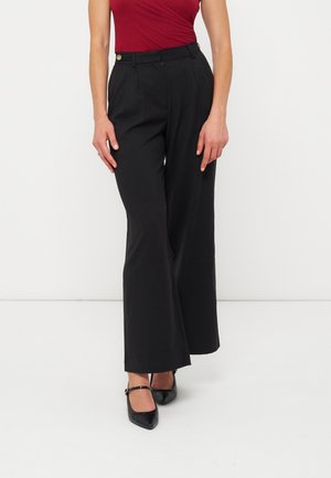 Femme portant un pantalon noir taille haute à jambes larges, un haut rouge sans manches et des talons Mary Jane noirs à bout pointu, debout sur un fond blanc.