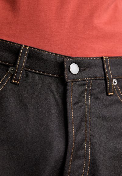 Black denim jeans with copper stitching, featuring a metal button closure and a textured finish, paired with a red shirt underneath.