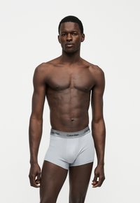 Μυώδης άντρας με σκουρόχρωμο δέρμα, που στέκεται με το πρόσωπο προς τα εμπρός φορώντας ανοιχτό γκρι εσώρουχα Calvin Klein μπροστά σε απλό λευκό φόντο.