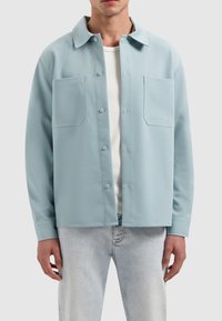 Lichtblauwe shirtjacket met een kraag, twee voorzakken, knoopsluiting en een gladde textuur, gedragen over een wit onderhemd.
