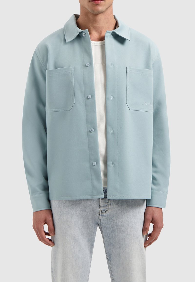 Lichtblauwe shirtjacket met een kraag, twee voorzakken, knoopsluiting en een gladde textuur, gedragen over een wit onderhemd.