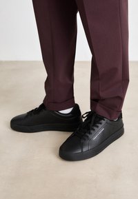Baskets noires en matériau lisse, dotées d'une toe arrondie, d'un système à lacets et d'un logo blanc sur le côté, assorties à un pantalon bordeaux.