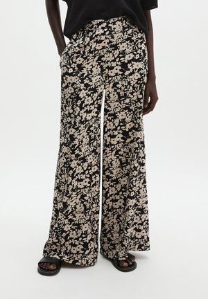 Femme portant un pantalon noir à jambes larges avec un motif floral beige et des sandales noires, debout les mains dans les poches.