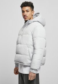 Hellegraue Pufferjacke mit Kapuze, die einen Reißverschluss vorne, elastische Manschetten und einen boxy Schnitt aufweist. Glatte, glänzende Textur.