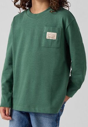 Child wearing a green long-sleeve shirt with a chest pocket and patch, paired with blue jeans, standing against a plain background.