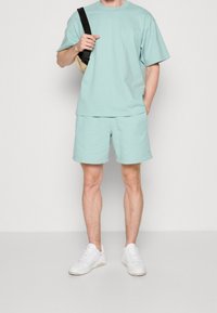 Minttu vihreä puuvillapaita ja shortsisetti, jossa on rento malli ja ribattu kaula-aukko, yhdistetty valkoisten lenkkareiden ja beigestä olkalaukusta.