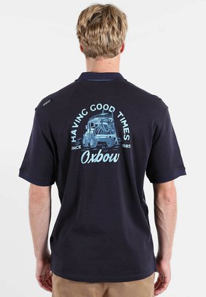 Homme blond aux cheveux courts portant un polo noir avec un graphique bleu clair d'un véhicule tout-terrain et le texte "Having Good Times Oxbow Since 1985"