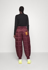 Weinrote Pufferhosen mit einer gesteppten Textur und elastischem Bund, ausgestattet mit einer seitlichen Taschen-Transporttasche mit einem braunen Etikett, kombiniert mit neongelben Turnschuhen.