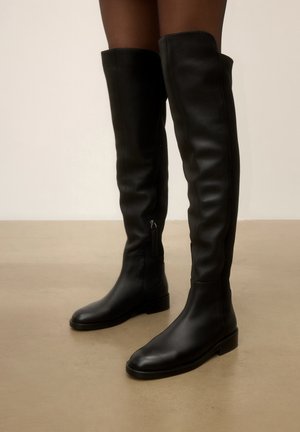 Bottes en cuir noires montant jusqu'au genou avec des talons bas portées par une personne debout sur un sol beige lisse.