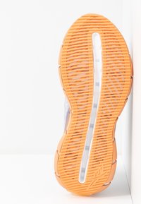 Semelle d'une basket avec une bande de roulement en caoutchouc orange et une section médiane transparente montrant le rembourrage interne, positionnée verticalement contre une surface blanche.