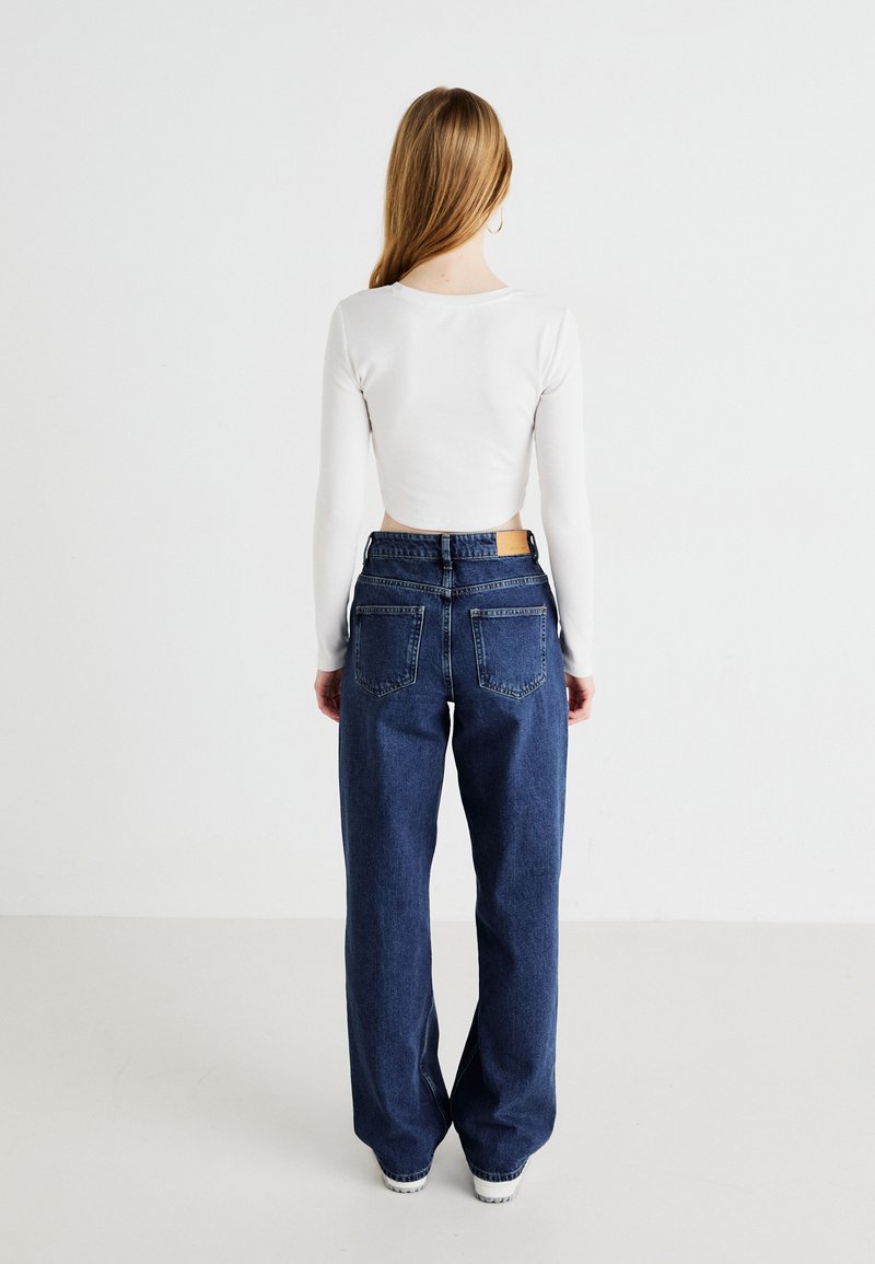 Biała, długa bluzka crop top z długim rękawem w zestawieniu z ciemnoniebieskimi, luźnymi dżinsami z wysokim stanem, które mają tylne kieszenie oraz brązową skórzaną naszywkę.