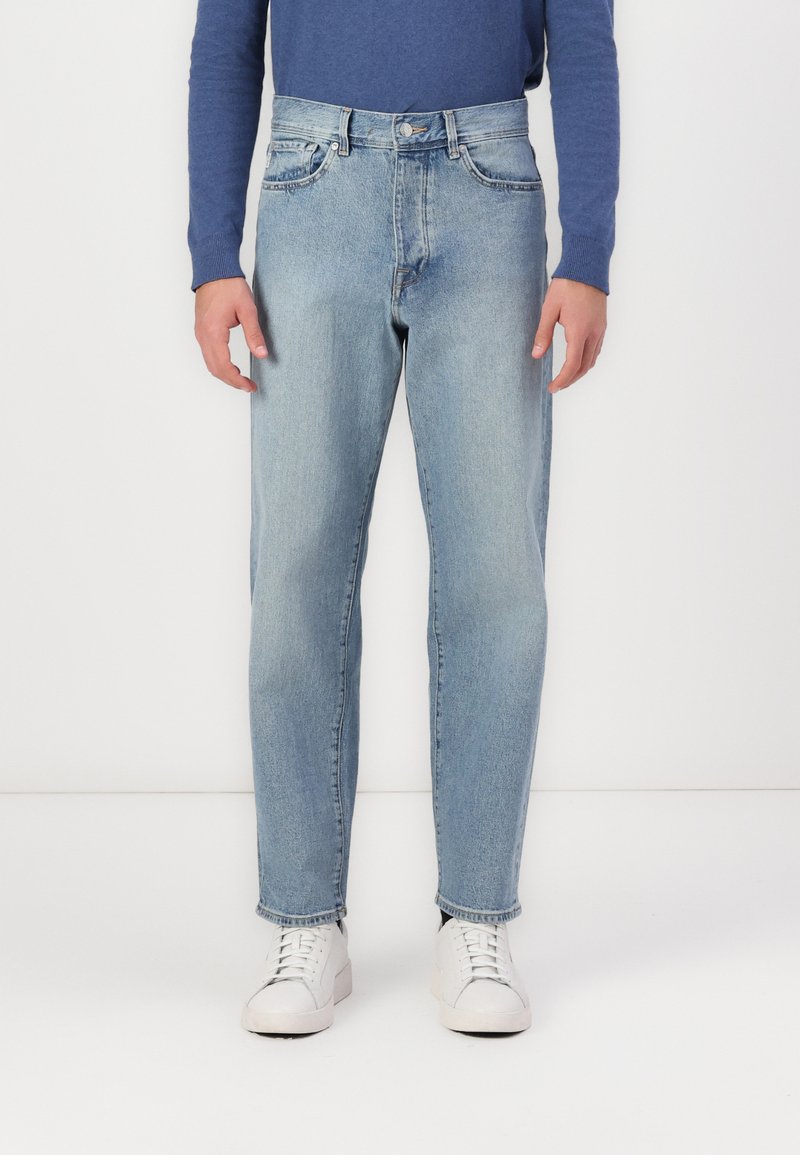 Jean en denim bleus clairs avec une coupe droite, comportant cinq poches, une fermeture à bouton et des coutures contrastées. Portés avec des baskets blanches.