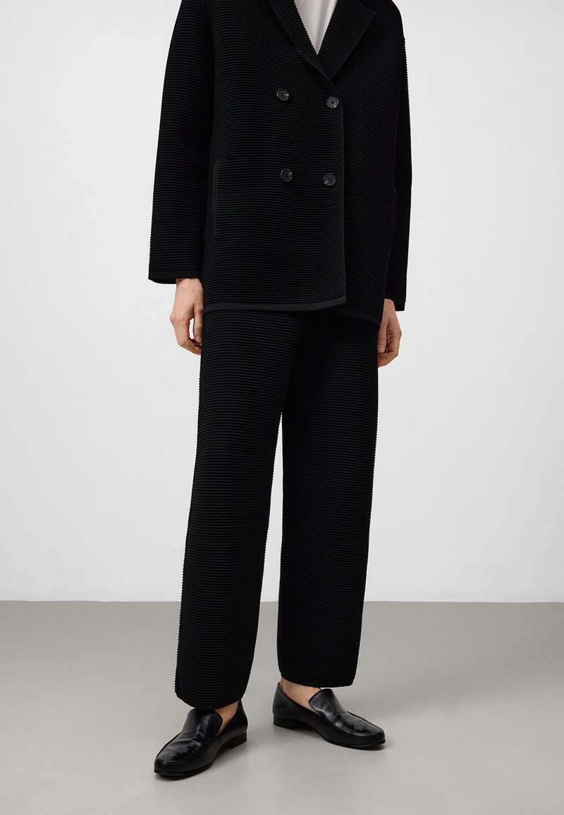 Giacca nera a costine doppiopetto con grandi bottoni, abbinata a pantaloni a costine coordinati. Indossata con scarpe nere slip-on.