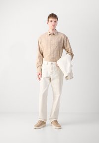 Svijetlosmeđi dijelovi s dugim rukavima, s suptilnim prugama, u kombinaciji s off-white hlačama i bež cipelama. Model drži odgovarajući sako.