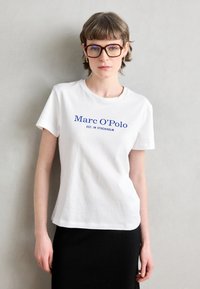 Balta kokvilnas T-kreklā ir zils teksts "Marc O'Polo EST. IN STOCKHOLM." Modelis valkā melnu ribotu svārku un brūnas taisnstūra brilles.