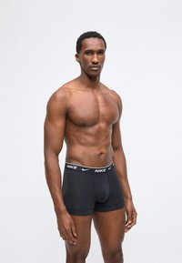 Muskuløs mann som står mot hvit bakgrunn, iført svarte Nike-boxershorts med synlig logo på linningen.