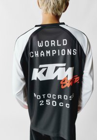 Čierno-biely dlhý rukáv dres s výrazným textom "SVETOVÍ ŠAMPIONI" a červeným logom "KTM". Vyznačuje sa športovým dizajnom a ľahkým materiálom.