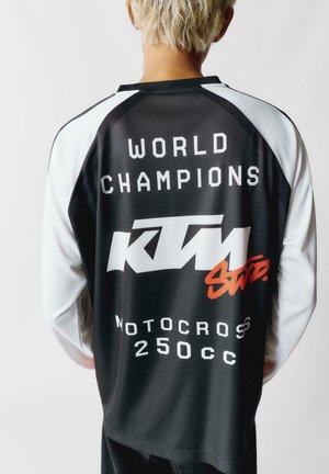 Čierno-biely dlhý rukáv dres s výrazným textom "SVETOVÍ ŠAMPIONI" a červeným logom "KTM". Vyznačuje sa športovým dizajnom a ľahkým materiálom.