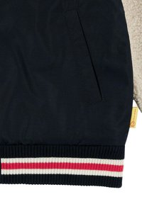 Tessuto esterno nero con un pannello in pile crema strutturato, caratterizzato da un orlo a costine con righe rosse e bianche. Include una tasca laterale.