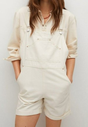 Kvinde iført cremefarvet langærmet skjorte under cremefarvede korte overaller med opkrøllede kanter og hænderne i lommerne.