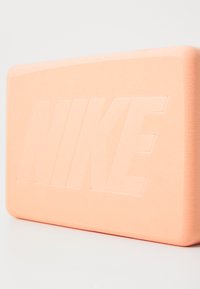 Sarkans Nike zīmola taisnstūra formas putu bloks maigi persiku krāsā, ar izspiestu logotipu vienā pusē, noapaļotiem stūriem un gludu tekstūru.