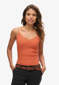 Top de tirantes naranja acanalado con borde de encaje, con tirantes finos. Combinado con un cinturón trenzado de cuero marrón y pantalones negros. Textura suave.