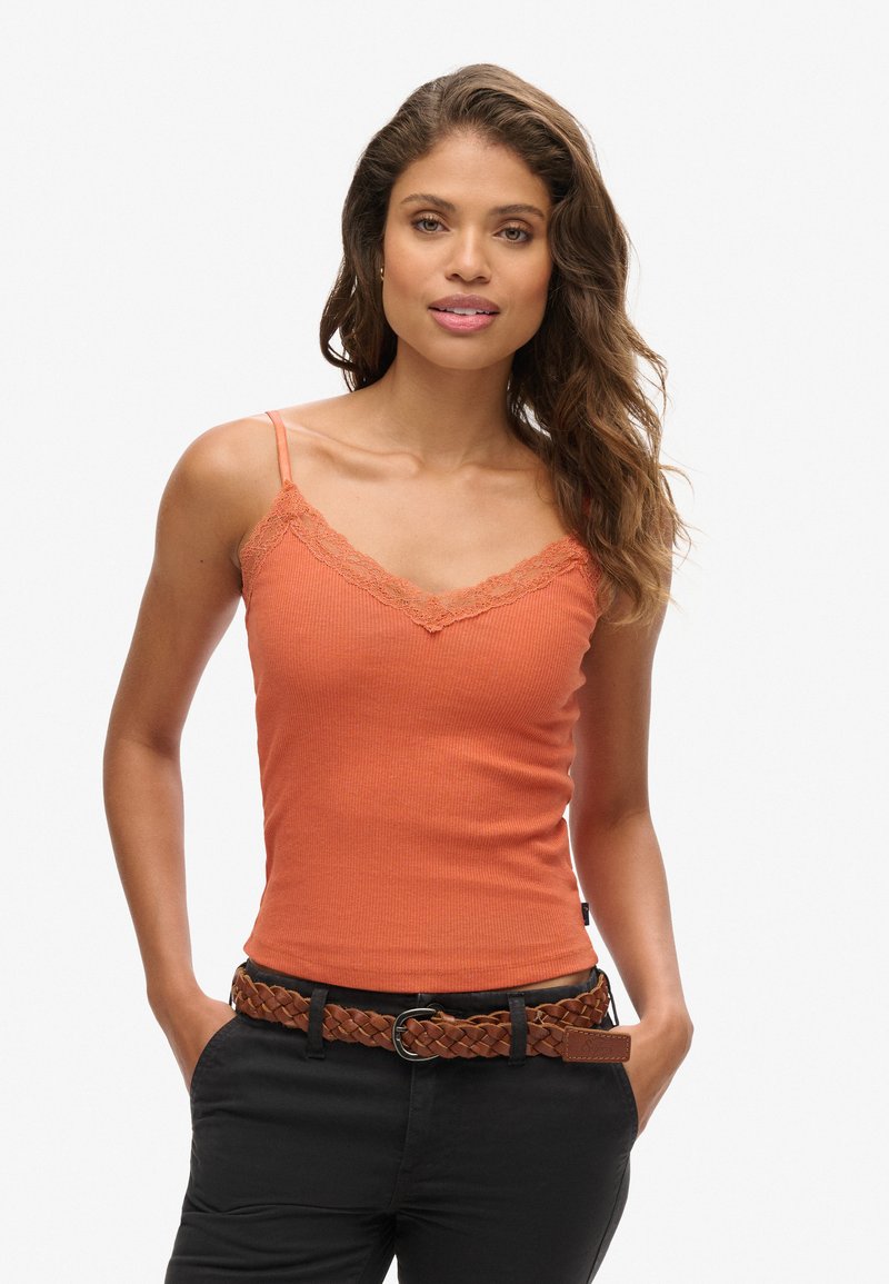 Top de tirantes naranja acanalado con borde de encaje, con tirantes finos. Combinado con un cinturón trenzado de cuero marrón y pantalones negros. Textura suave.