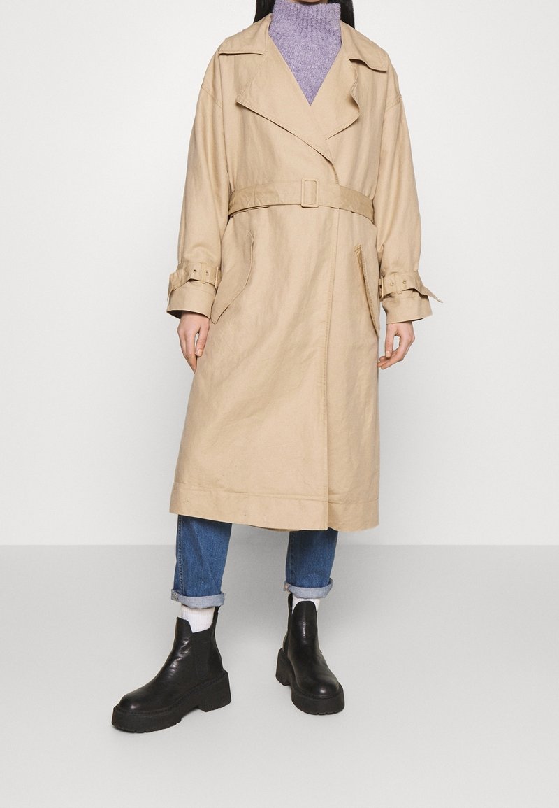 Personne portant un trench-coat beige ceinturé par-dessus un col roulé violet, un jean bleu retroussé, des chaussettes blanches et des bottines noires épaisses.