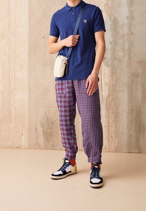 Person trägt ein marineblaues Poloshirt, rot-blau kariere Hose, mehrfarbige High-Top-Sneaker und eine kleine beige Umhängetasche.