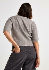 Kortærmet, lysegrå strikket sweater med ribbet kant. Sammen med mørkegrå løstsiddende jeans med baglommer. Set fra ryggen.