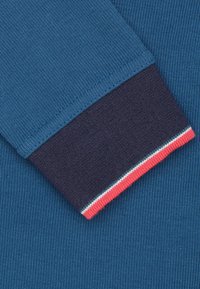 Lichtblauwe sweaterstof met ribbelstructuur, met een donkerblauwe manchette met rode en witte streepaccenten. Close-up van de details.