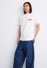 White cotton t-shirt with short sleeves, featuring a small blue graphic on the left chest. Paired with loose-fit blue denim pants.