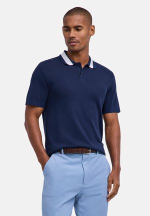 Uomo che indossa una polo blu navy a maniche corte con colletto a righe bianche e pantaloni azzurri con cintura intrecciata marrone.