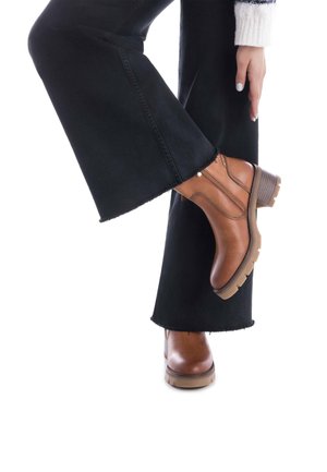 Botas de tobillo de cuero marrón con cremalleras laterales, un tacón apilado y una suela de goma texturizada, combinadas con jeans negros de pierna ancha.