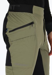 Olijfgroene wandelbroeken met zwarte accenten, taps toelopende pasvorm, contrasterend stofpaneel, ritssluitingzak en logo aan de zijkant.