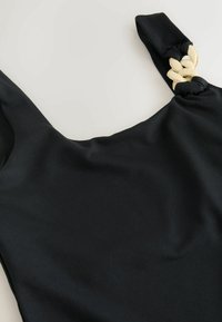 Haut noir sans manches avec un col carré et un détail beige tressé sur une bretelle, présenté sur un fond clair.