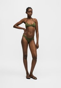 Femme debout, une main sur la hanche, portant un bikini vert olive et des tongs noires sur un fond clair uni.
