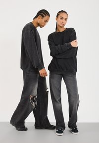 Sorte overdimensjonerte hettegensere og slitte sorte jeans på to modeller. Stoffet ser mykt ut, med en falmet tekstur og synlige hull på jeansene.