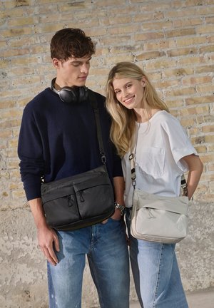 Young man and woman stand against a brick wall, wearing casual clothes with crossbody bags; he wears headphones around his neck.