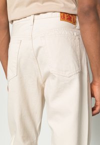 Pantalones de mezclilla beige con pierna recta, que cuentan con un bolsillo trasero, una textura sutil y una etiqueta tejida roja en la cinturilla.