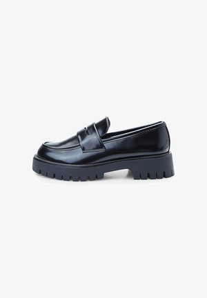 Musta lakattu nahka loafers, joissa on paksu harmaa kumipohja, koristeellinen hihna ja selkeä ommel reunoilla.