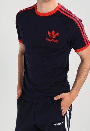 T-shirt de sport bleu marine avec des accents rouges. Caractéristiques : col côtelé rouge, trois bandes rouges sur les épaules, et logo Adidas rouge.