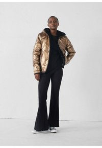 Persona vistiendo una chaqueta metálica bronce sobre una camiseta negra, pantalones negros acampanados y zapatillas blancas y negras, de pie contra una pared blanca lisa.