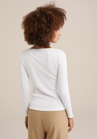 Top blanco de manga larga acanalada con escote barco, silueta ajustada y textura suave, combinado con pantalones beige claros.