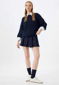 Maglione spesso a trecce blu navy sopra una camicia bianca con colletto, abbinato a una gonna plissettata blu navy. Il modello indossa sneakers bianche e calzini blu navy.