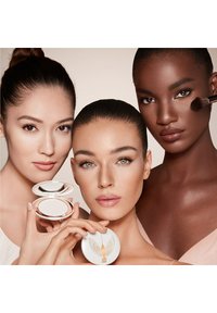 Three women with various skin tones feature a compact makeup product. The packaging is white with a gold decorative logo and wings.