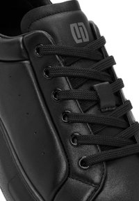 Zapatilla negra con parte superior de cuero suave, cordones texturizados, acompañada de ojetes de metal, con un logo en la lengüeta y un diseño minimalista.